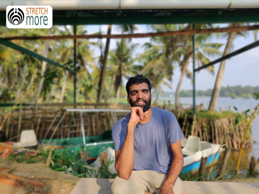 Outdoor, Nibin is sitting on a bench. His chin is leaning on his right hand while he looks directly at the camera. He wears a light blue shirt and light gray pants. Behind Nibin there are boats but since it's portrait, they are blur. Also on the top left of the image shows the logo of Stretch More.